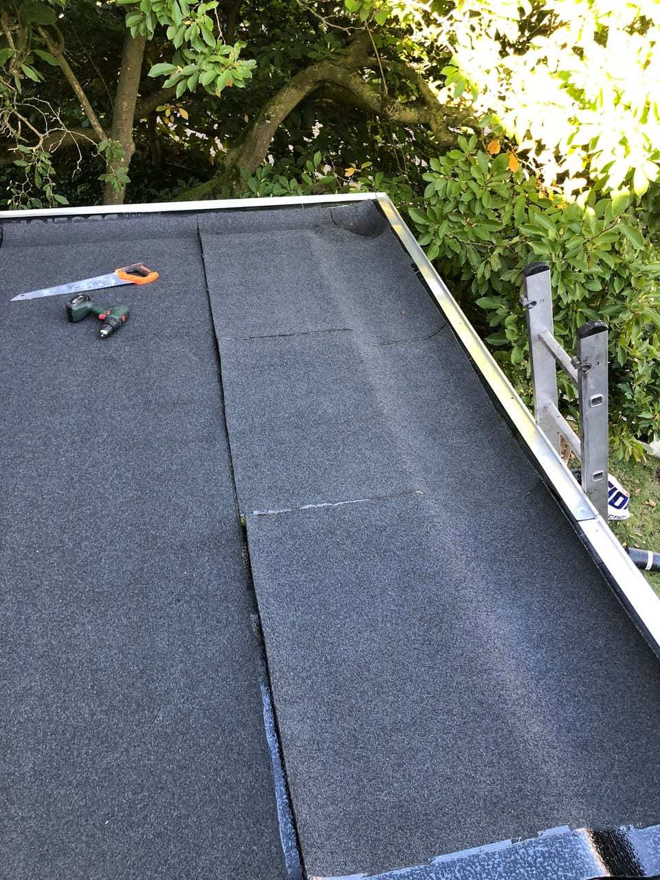 Nieuw bitumen dak met een aluminium daktrim, een zaag en een boormachine, met een ladder tegen de dakrand