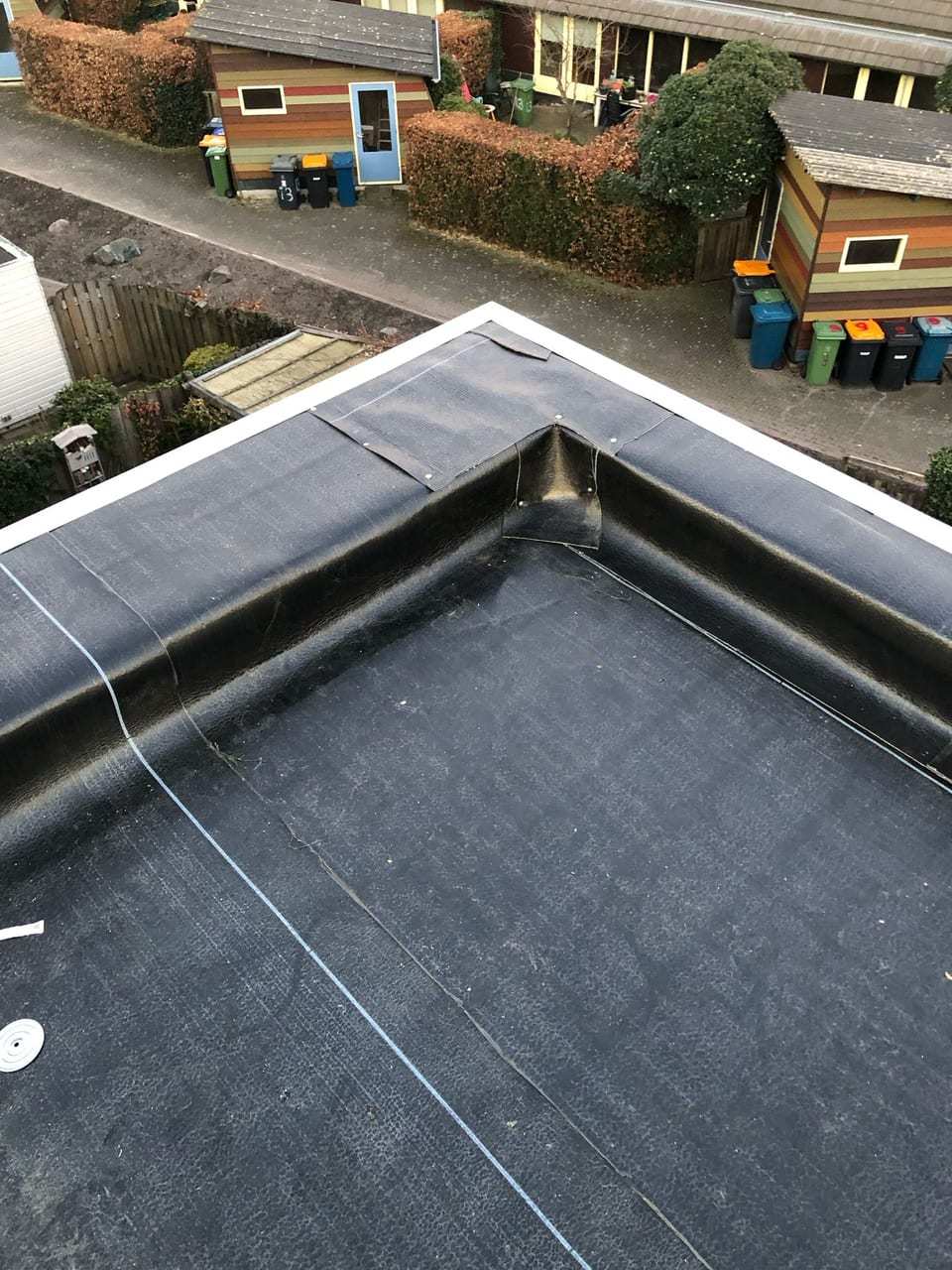 Hoek van een plat dak met nieuwe bitumen en een opstaande dakrand, boven achtertuinen met schuurtjes