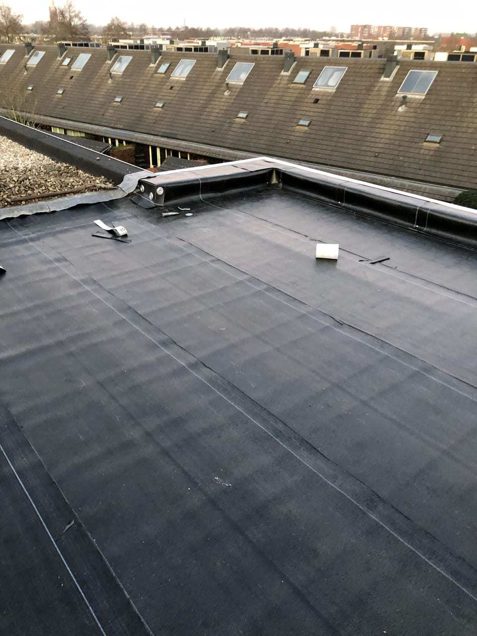 Plat dak met nieuwe zwarte bitumen, met daarachter een rij woningen met pannendaken en dakramen