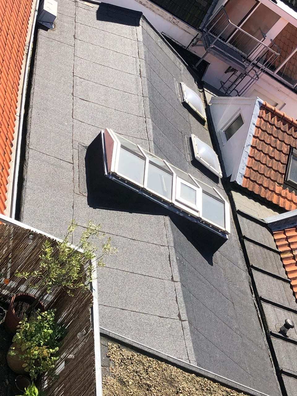 Plat dak met bitumen en een lichtstraat, van bovenaf gezien, tussen daken met oranje pannen