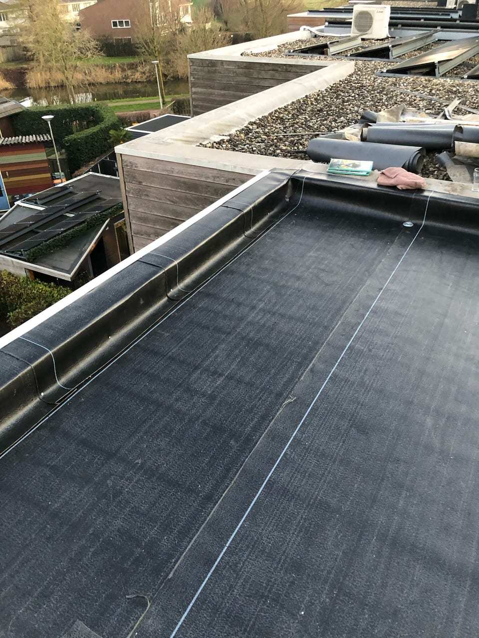 Plat dak met nieuwe bitumen en een opstaande rand met daktrim, naast een plat dak met grind